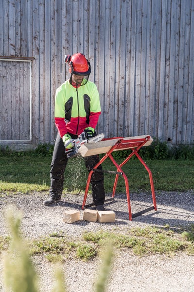 Eine Person in Schutzausrüstung sägt mit einer Akku-Kettensäge einen Holzbalken auf einem roten Sägebock aus Metall im Freien.