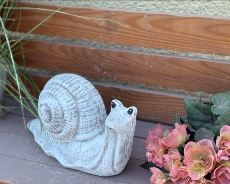 Dekorative Gartenfigur in Schneckenform aus wetterfestem Steinmaterial auf einer Holzbank mit Blumen und Gras.