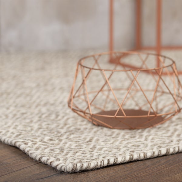 Handgewebter Teppich mit geometrischem Rautenmuster in Beige und Braun auf dunklem Holzboden mit einem Dekokorb aus Draht.