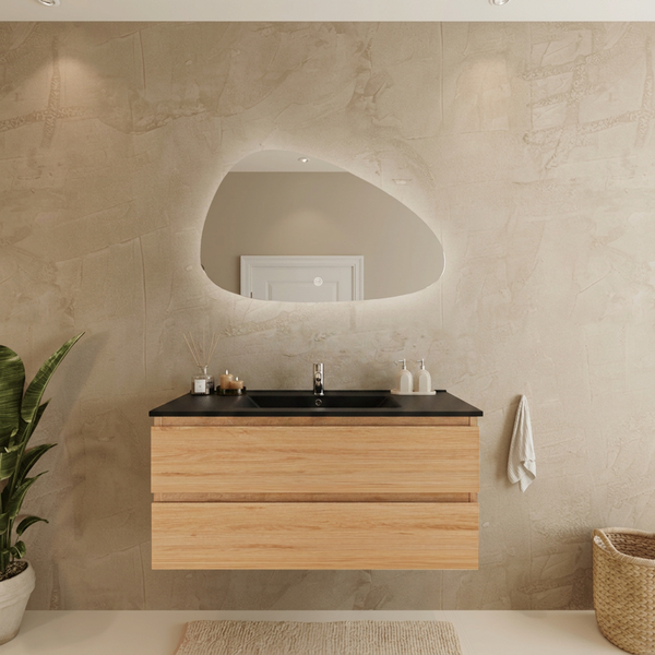 Moderner Badezimmer-Waschtisch in Holzoptik mit schwarzem Waschbecken und einem asymmetrisch geformten, beleuchteten Wandspiegel.