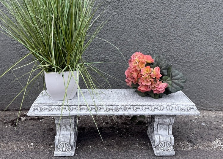 Kleine Steingut-Gartenbank mit dekorativen Verzierungen, auf der ein Pflanztopf mit Gras und künstliche Blumen stehen.