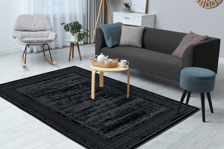 Moderner Wohnbereich mit dunklem Teppich mit Ornament-Bordüre, grauem Sofa, Schaukelstuhl und hellem Holztisch auf hellem Boden.