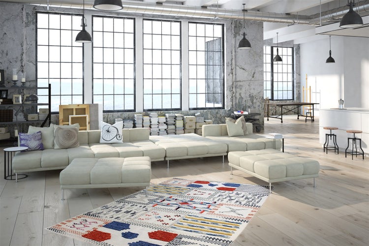 Helles, geräumiges Loft-Wohnzimmer mit großem modularem Sofa, bunt gemustertem Teppich und Industrie-Fenstern vor Betonwänden.