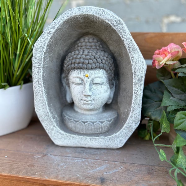 Buddha-Kopf in einer ovalen Steinnische, dekorative Gartenfigur aus Steinguss, platziert auf einem Holzregal neben Pflanzen.