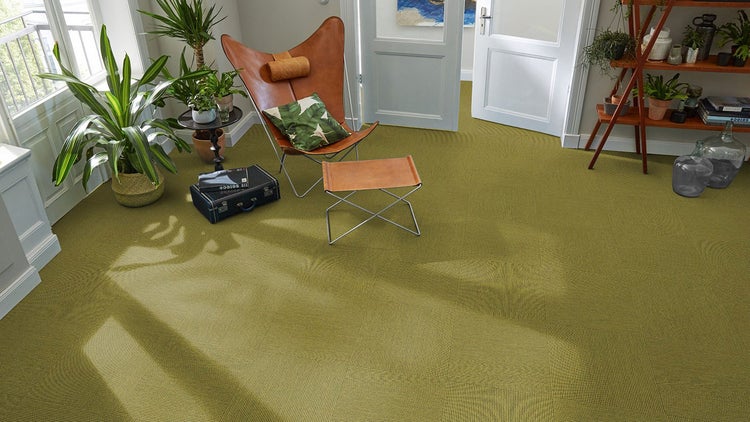 Helles Wohnzimmer mit grünen, strukturierten Teppichfliesen, einem Butterfly-Sessel aus Leder mit Hocker und verschiedenen Zimmerpflanzen.