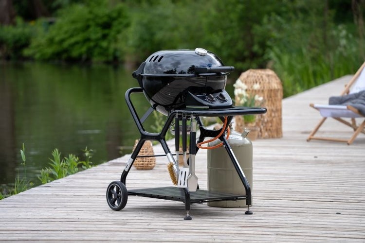 Schwarzer Gasgrill auf Holzsteg am Wasser mit Grillbesteck und Gasflasche. Organisierte Außenszene mit Thermometer und seitlicher Ablagefläche.