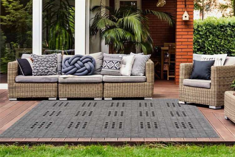Rechteckiger Outdoorteppich mit grau-schwarzem Muster auf einer Holzterrasse mit Polyrattan-Gartenmöbeln, Dekokissen und grünen Pflanzen.