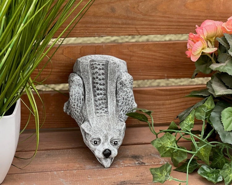 Gartenfigur aus Steinguss in Form eines kletternden Fabelwesens, aufgehängt an einem Holzzaun neben Pflanzen.