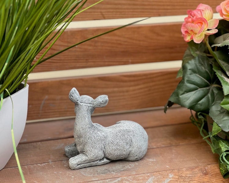 Kleine Gartenfigur liegendes Rehkitz in Steinoptik auf einer Holzterrasse zwischen zwei Topfpflanzen.