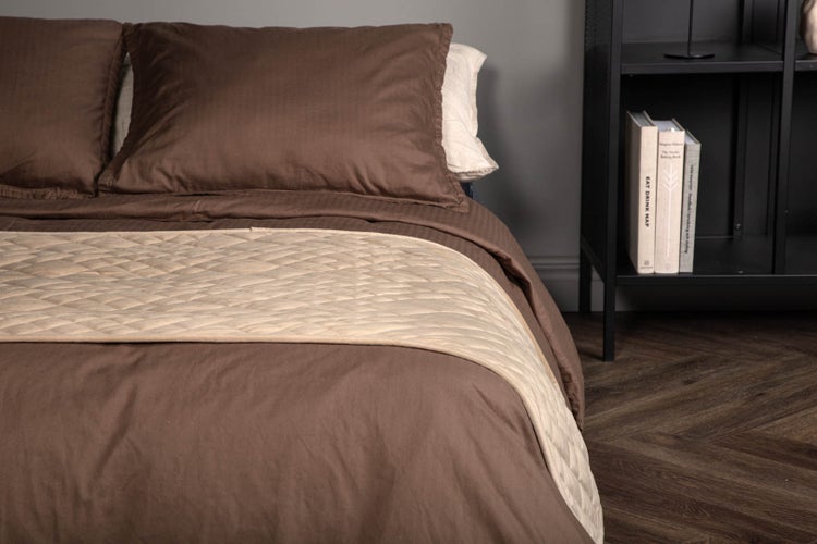 Bett mit brauner Bettwäsche und gestepptem beigefarbenem Bettläufer neben einem schwarzen Metallregal auf Holzboden.