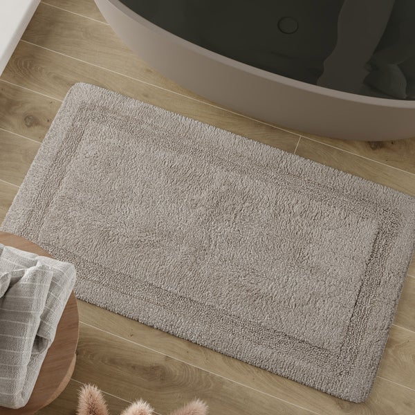 Rechteckige Badematte mit Bordüren-Design auf Boden in Holzoptik neben einer Badewanne. Weiches Material mit Schlingenstruktur.