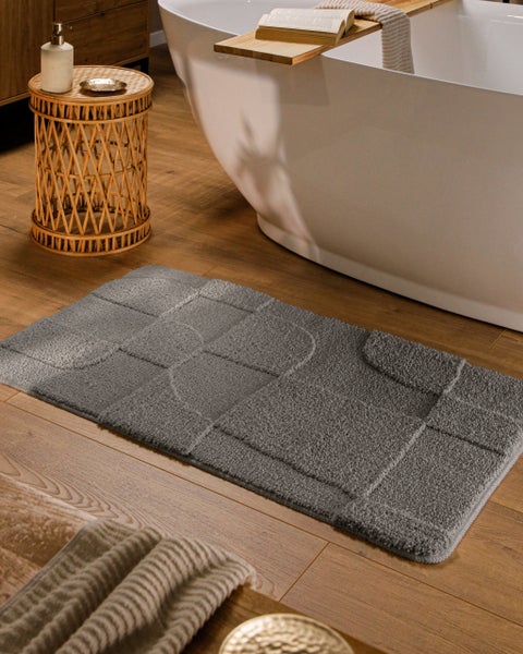 Graue Badematte mit geometrischem Muster vor weißer freistehender Badewanne in hellem Badezimmer mit Holzoptik-Boden.