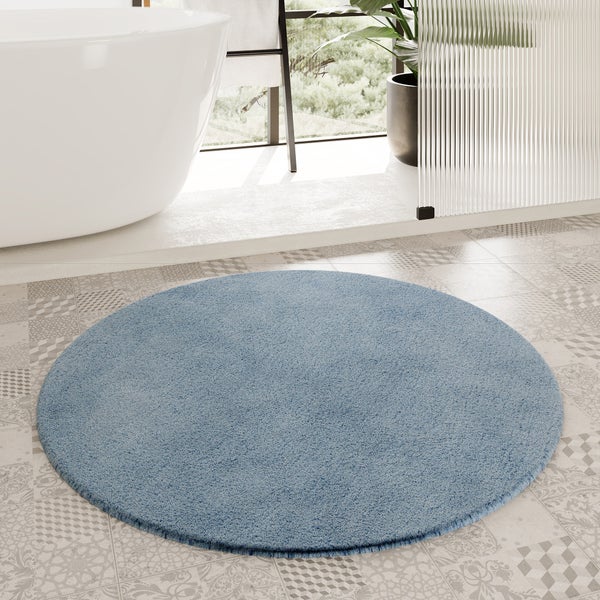 Runder blauer Badteppich in einem hellen Badezimmer mit freistehender Badewanne, geriffeltem Glas und gemusterten Bodenfliesen.