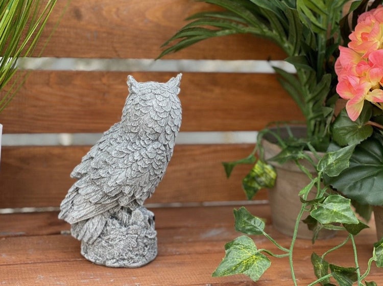 Dekorative Eulenfigur aus Steinoptik zur Gartendekoration auf einem Holzuntergrund neben einer Pflanze.
