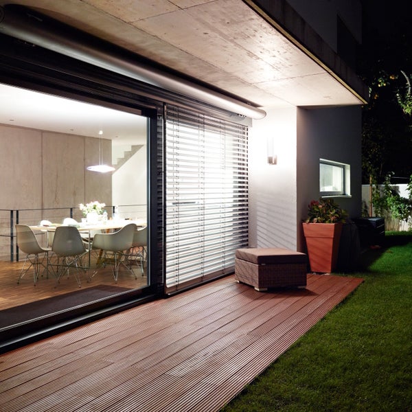 Moderne Terrasse mit Holzbelag vor einem Haus mit bodentiefen Fenstern und Außenjalousien bei Nacht.