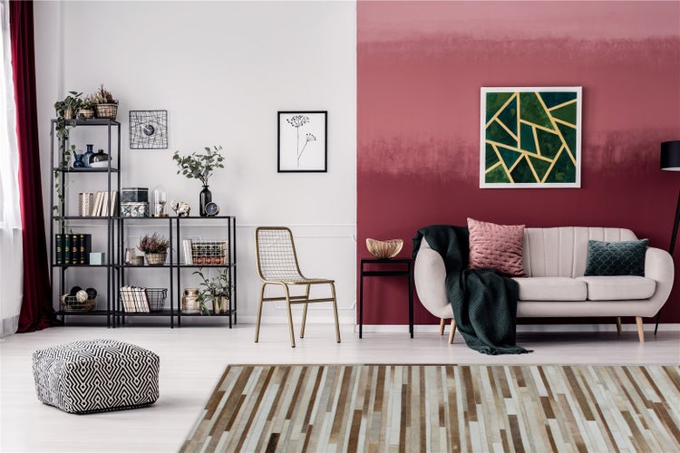 Modernes Wohnzimmer mit beigem Sofa, Wand mit rotem Farbverlauf, schwarzem Metallregal, goldenem Stuhl und gestreiftem Teppich in Brauntönen.