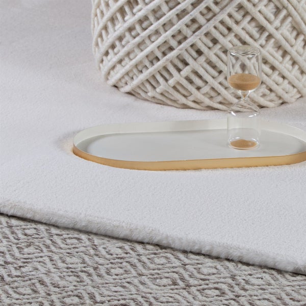 Weicher weißer Teppich in Felloptik auf einem gemusterten Teppich mit einem Dekutablett, einer Sanduhr und einem grob gestrickten Korb.