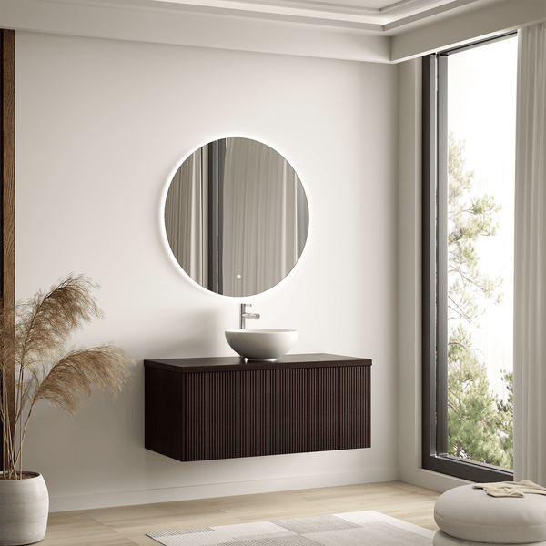 Modernes Badezimmer mit dunklem Waschtischunterschrank, rundem Aufsatzwaschbecken und beleuchtetem Rundspiegel an einer hellen Wand.