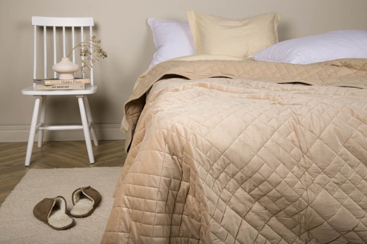 Gesteppte beige Tagesdecke auf einem Bett im Schlafzimmer neben einem weißen Stuhl mit Dekoration und Teppich mit Hausschuhen.