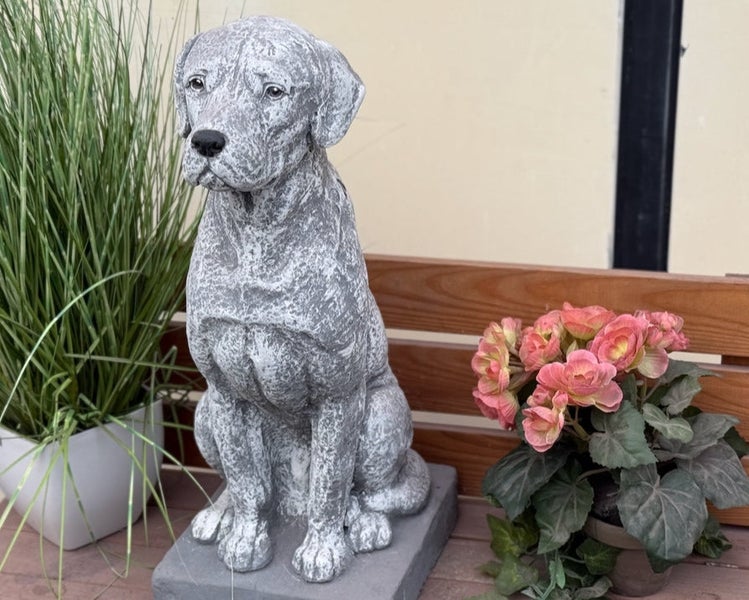 Sitzende Hundefigur aus Steinguss im Labrador-Stil, dekorative Gartenfigur neben Topfpflanzen auf einer Holzbank.