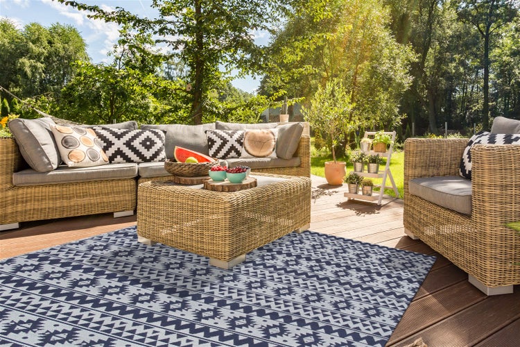 Modernes Garten-Lounge-Set aus Geflecht mit Ecksofa, Sessel, Tisch und gemustertem Outdoor-Teppich auf einer hellen Holzterrasse im Garten.