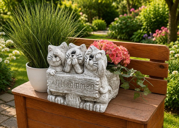 Steinfigur drei Katzen mit Willkommen Schild als Pflanzgefäß auf einer Gartenbank mit Topfpflanze und Hortensienblüten im Hintergrund.