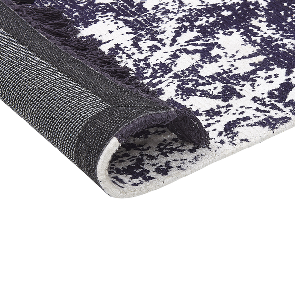 Detail eines Teppichs mit abstraktem, gesprenkeltem Muster und dunklen Fransen. Die umgeklappte Ecke zeigt die strukturierte Rückseite.