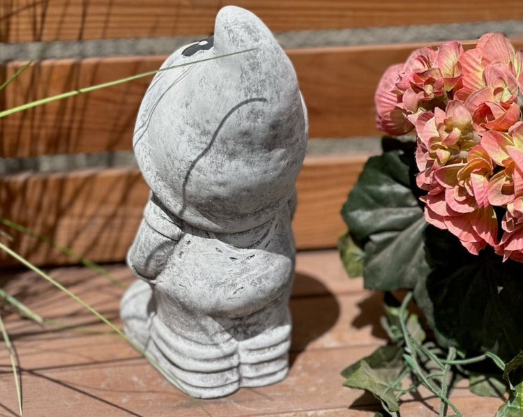 Gartenzwerg in Steinoptik von hinten auf einer Holzoberfläche stehend, neben einer Pflanze mit Blüten.