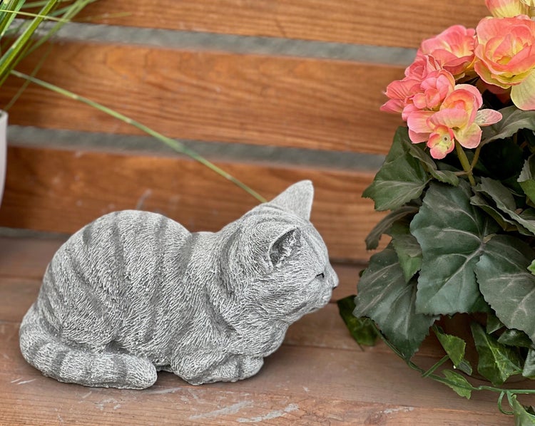 Gartenfigur in Form einer liegenden, getigerten Katze in Steinoptik, platziert neben einer Topfpflanze.