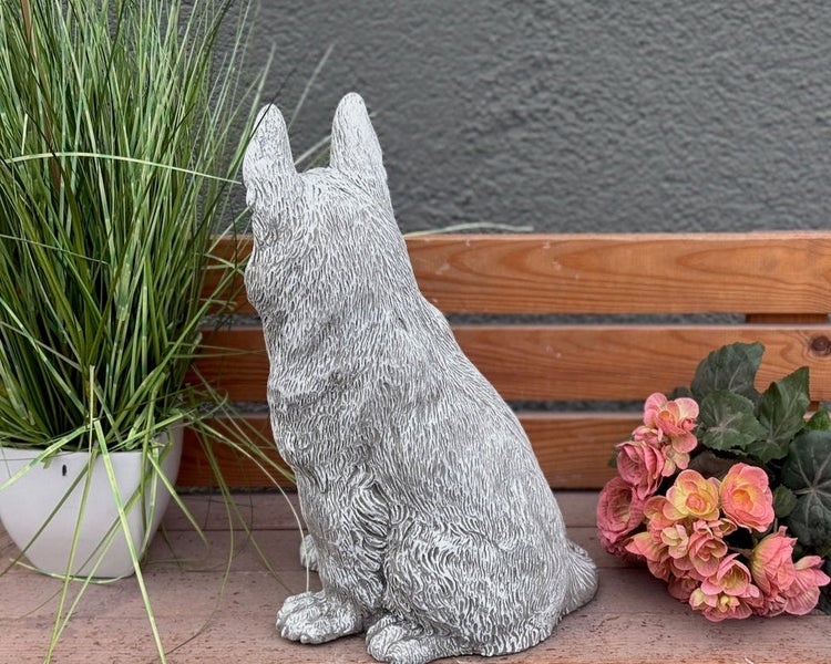 Gartendekoration Hundefigur aus Steinguss, sitzende Hundefigur von hinten auf einer Holzbank neben Pflanzen.