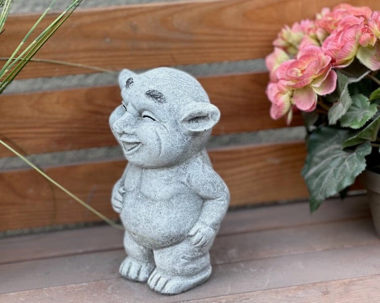 Steinerne Gartenfigur in Form eines lachenden Trolls stehend auf einer Holzfläche.