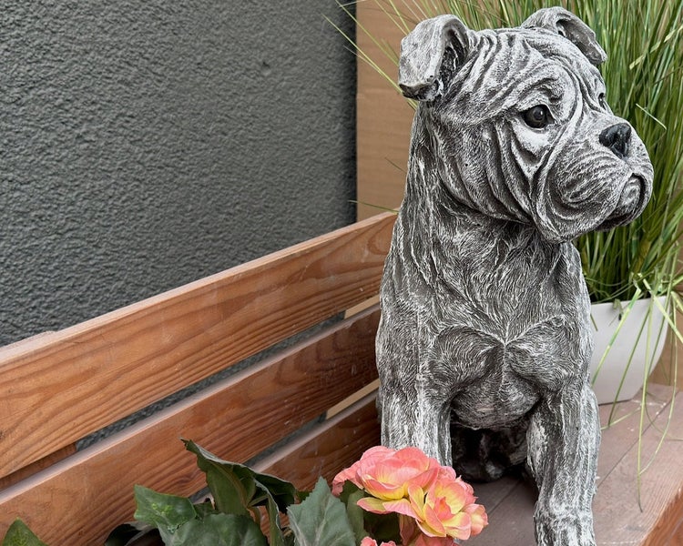 Detailreiche Gartenfigur eines sitzenden Hundes aus Steinguss auf einer Holzbank neben Blumen.