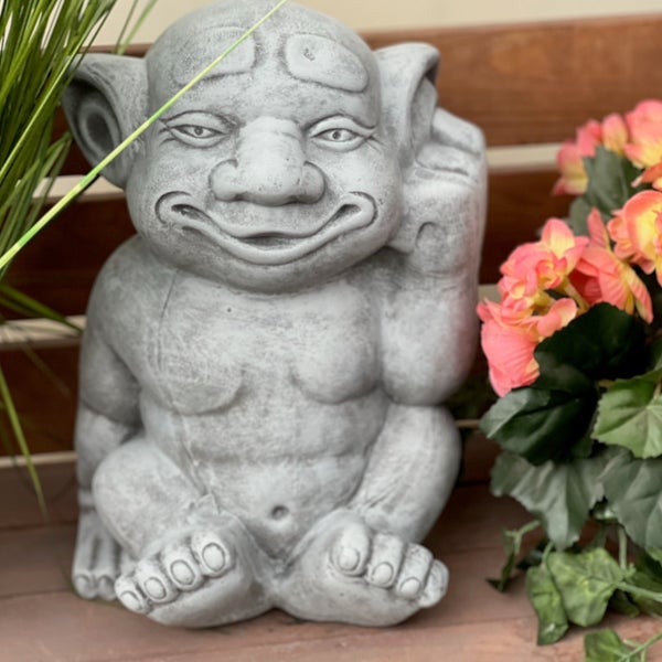 Steinfigur Troll aus wetterfestem Material zur Dekoration im Garten auf einem Holzuntergrund mit Blumen und Gras.