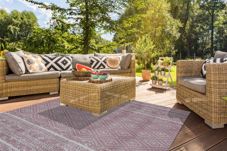 Hell gestaltete Terrasse mit Geflecht-Gartenmöbeln und einem lila Outdoorteppich mit geometrischem Muster auf Holzdielen im Garten.