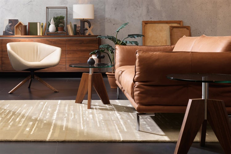 Modernes Wohnzimmer mit braunem Ledersofa, cremefarbenem Drehsessel, Sideboard und Beistelltischen auf gemustertem Teppich. Hell und geordnet.