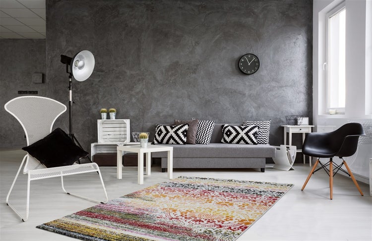 Moderner Wohnraum mit grauer Wand in Betonoptik, grauem Sofa, buntem Teppich mit Muster und verschiedenen Designstühlen.