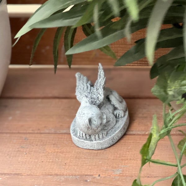 Kleine Grabfigur eines liegenden Engels aus Stein auf einer Holzunterlage mit Pflanzen im Hintergrund