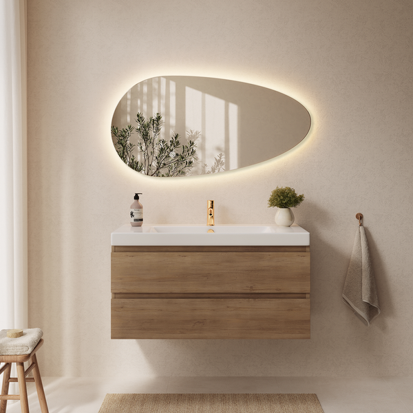 Badezimmer mit organisch geformtem Wandspiegel mit LED Beleuchtung, hängendem Waschunterschrank in Holzoptik und goldfarbener Waschtischarmatur.