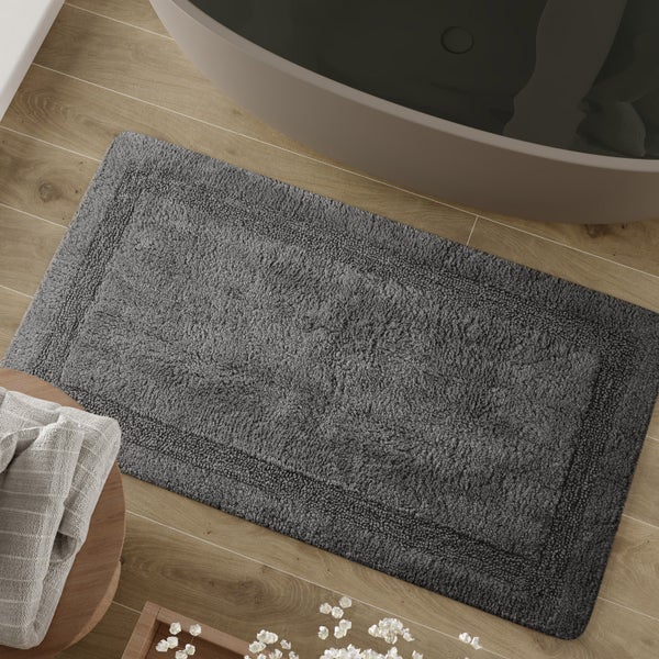 Graue Badematte mit Rahmenstruktur aus Baumwolle auf einem Holzfußboden neben einer Badewanne.