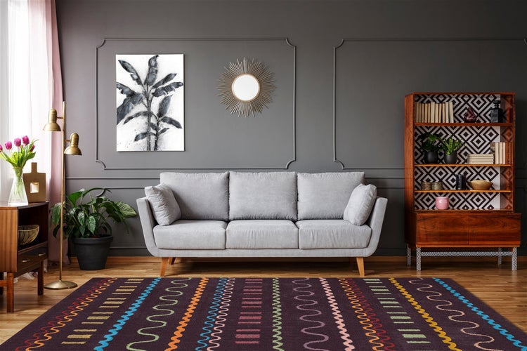 Modernes Wohnzimmer mit grauem Sofa, gemustertem lila Teppich, Holzregal und Sonnenspiegel an einer dunkelgrauen Wand mit Wandleisten.