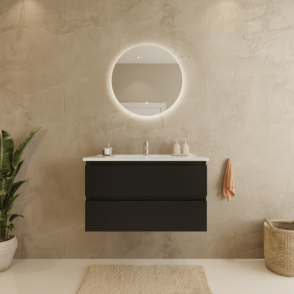 Modernes Badezimmer mit dunkelgrauem Hängewaschbeckenunterschrank, weißem Waschtisch und rundem, beleuchtetem Spiegel vor einer beigen Wand.