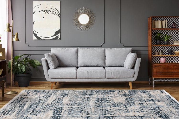 Modernes Wohnzimmer mit grauem Dreisitzer-Sofa, gemustertem Teppich, Holzregal, goldener Stehlampe und Sonnenspiegel vor grauer Wand.