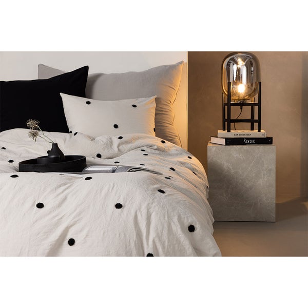 Schlafzimmer-Szene mit Bettwäsche mit Pompons, verschiedenen Kissen und einem modernen Nachttisch in Steinoptik mit einer Tischleuchte.