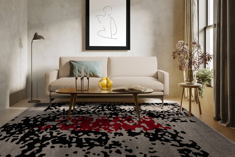 Moderner Teppich mit abstraktem Muster in Grau, Schwarz und Rot in einem hellen Wohnzimmer mit beigem Sofa und Holztisch.