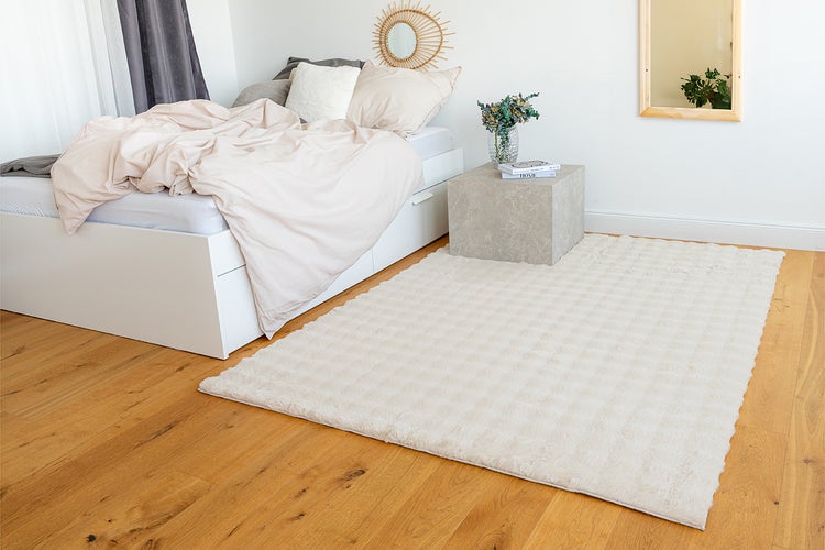 Ein flauschiger, cremefarbener Teppich in einem hellen Schlafzimmer mit weißem Bett und Holzfußboden.