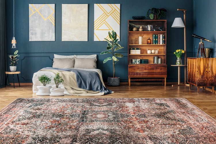 Modernes Schlafzimmer mit dunkelblauer Wand, großem gemustertem Teppich, Bett, Bücherregal aus Holz, Zimmerpflanzen und abstrakter Wandkunst.