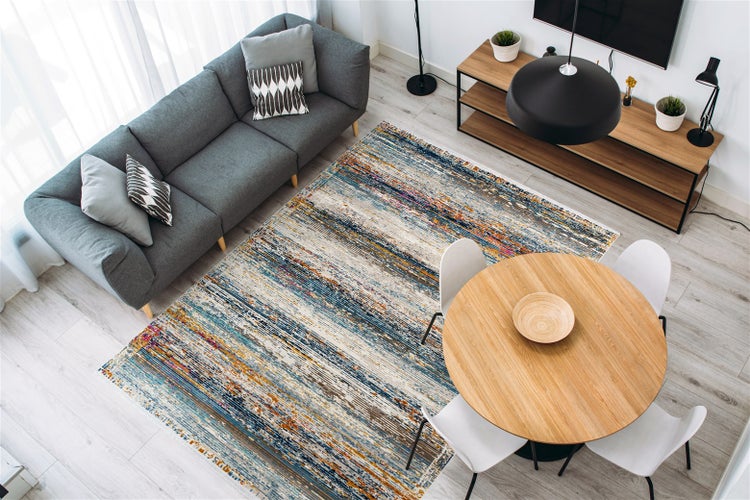 Modernes helles Wohnzimmer mit buntem Teppich im abstrakten Streifenmuster, grauem Sofa, Holzmöbeln und weißen Stühlen.