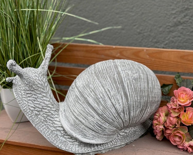 Gartenfigur Schnecke in Steinoptik mit strukturierter Oberfläche auf einer Holzbank neben Pflanzen.