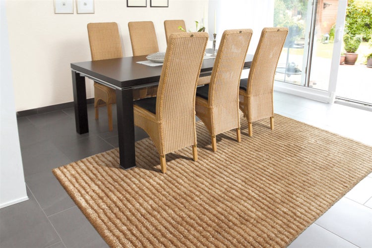 Hell gestaltetes Esszimmer mit einem dunkelbraunen Tisch, Rattanstühlen und einem strukturierten, gestreiften Teppich auf dunklen Bodenfliesen.