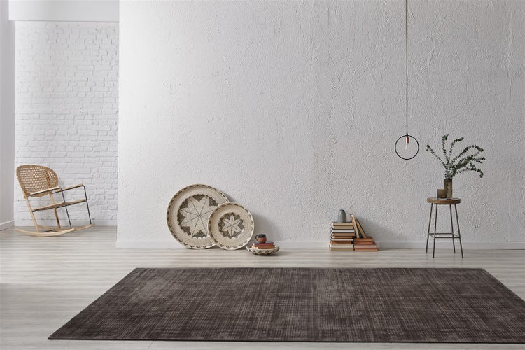 Moderner Wohnraum mit einem dunklen, strukturierten Teppich auf hellem Holzboden, weißen Wänden, einem Schaukelstuhl und minimalistischer Deko.